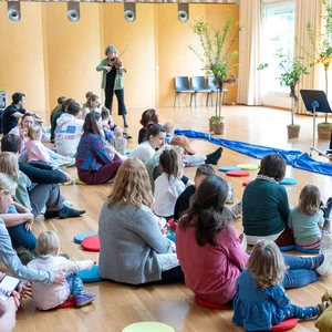 Eine Gruppe von Erwachsenen und Kindern sitzt auf dem Boden in einem Raum und hört zwei Musikerinnen, die Geige und Cello spielen.