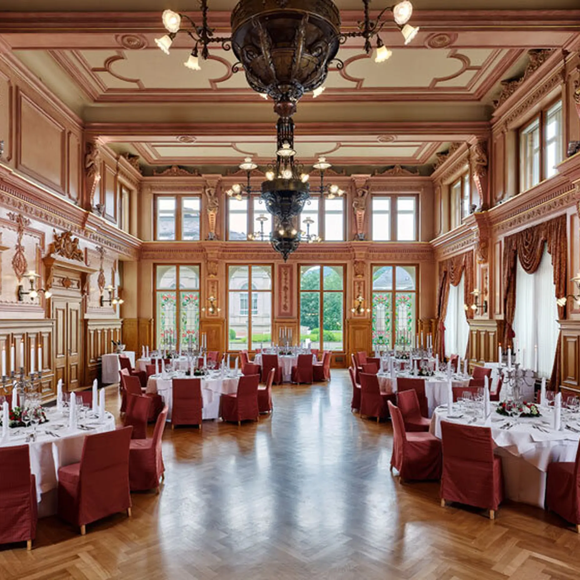An elegant, large banquet hall with set tables, red chairs, and ornate wood paneling. Chandeliers and large windows adorn the room.