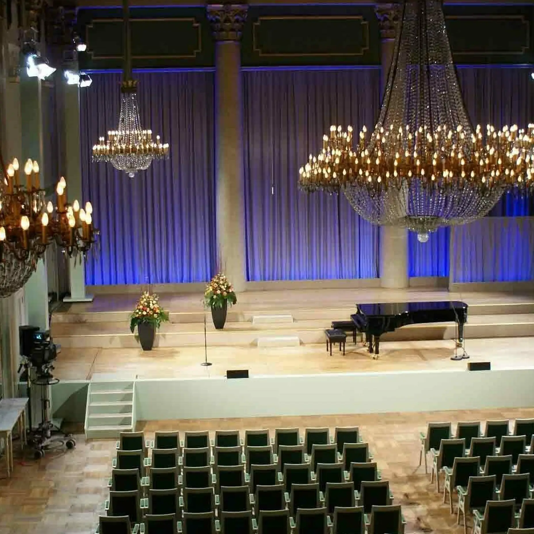 An elegant concert hall with chandeliers, a stage with a grand piano, and empty seating. Subdued lighting creates a serene atmosphere.