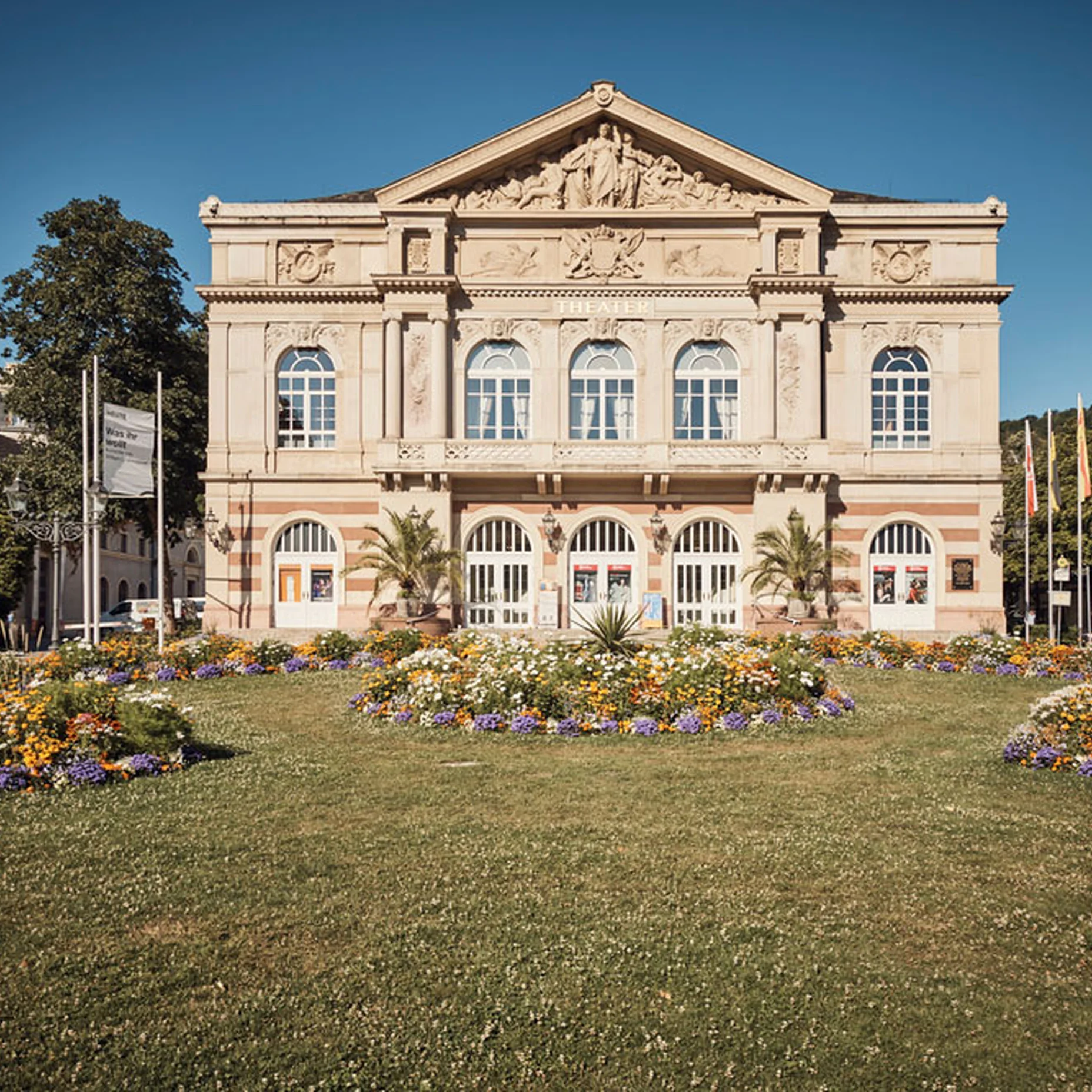 Historisches Gebäude mit klassischer Fassade, flankiert von Bäumen und Blumenbeeten im Garten. Blauer Himmel als Hintergrund.